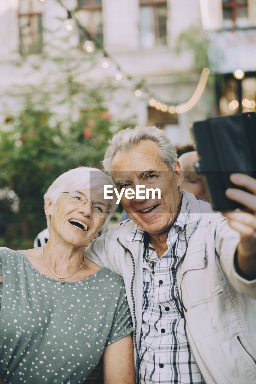 Senior man taking selfie with happy woman while sitting at restaurant in city