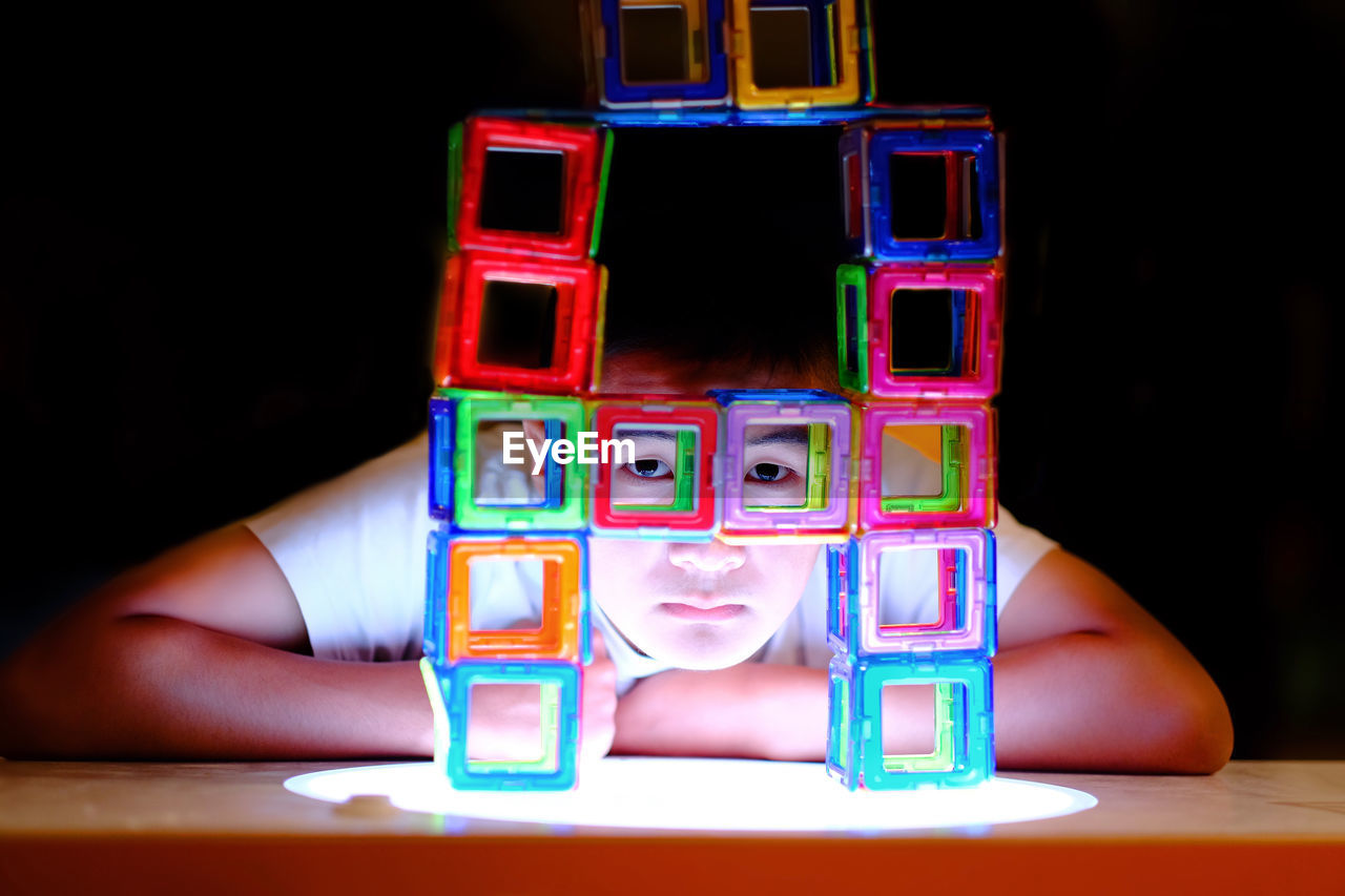 Portrait of boy seen through multi colored toy blocks by table against black background