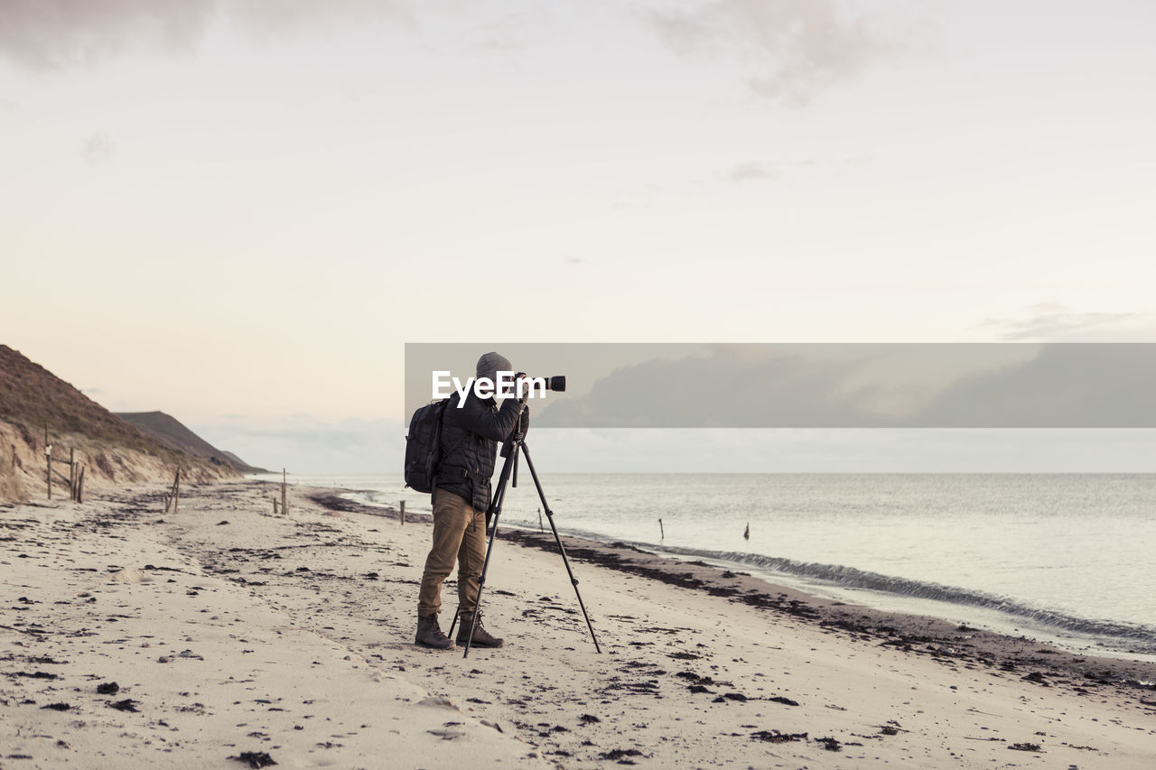 Full length side view of hiker photographing through slr camera on shore at beach