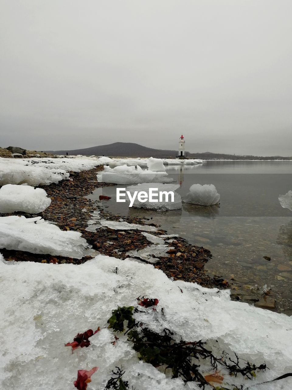 View of seashore with ice floes and a lighthouse in the distance during foggy spring morning