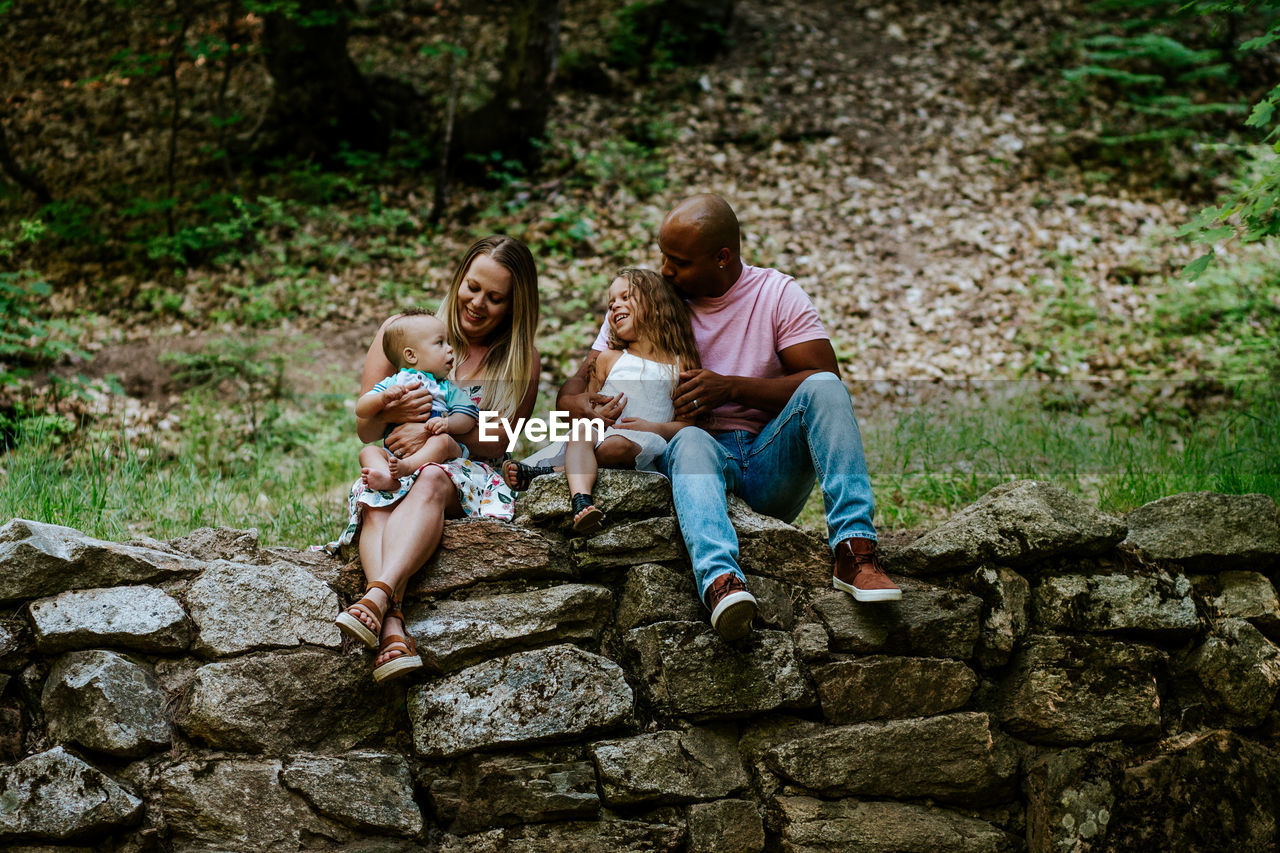 Happy young diverse family sitting on rocks in forest smiling