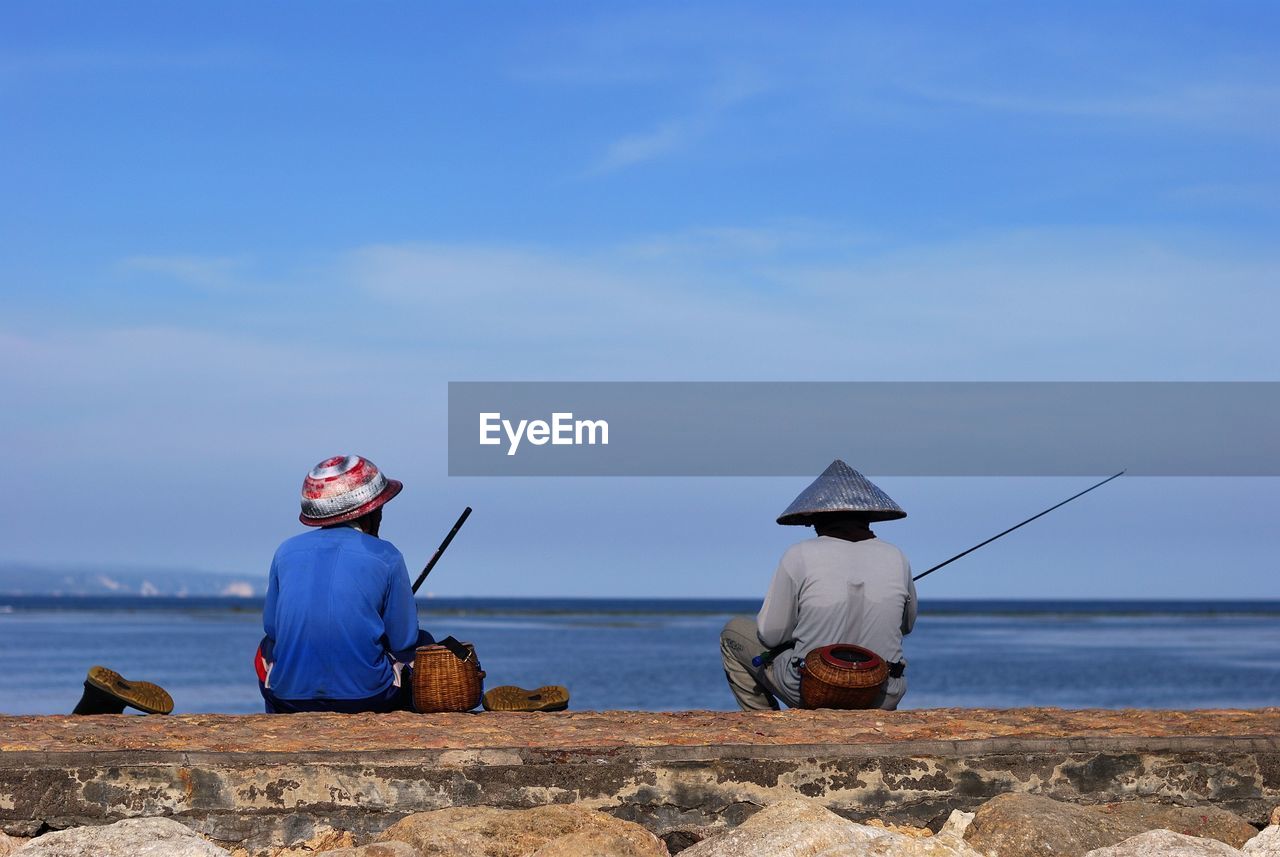 Rear view of men fishing by lake against sky