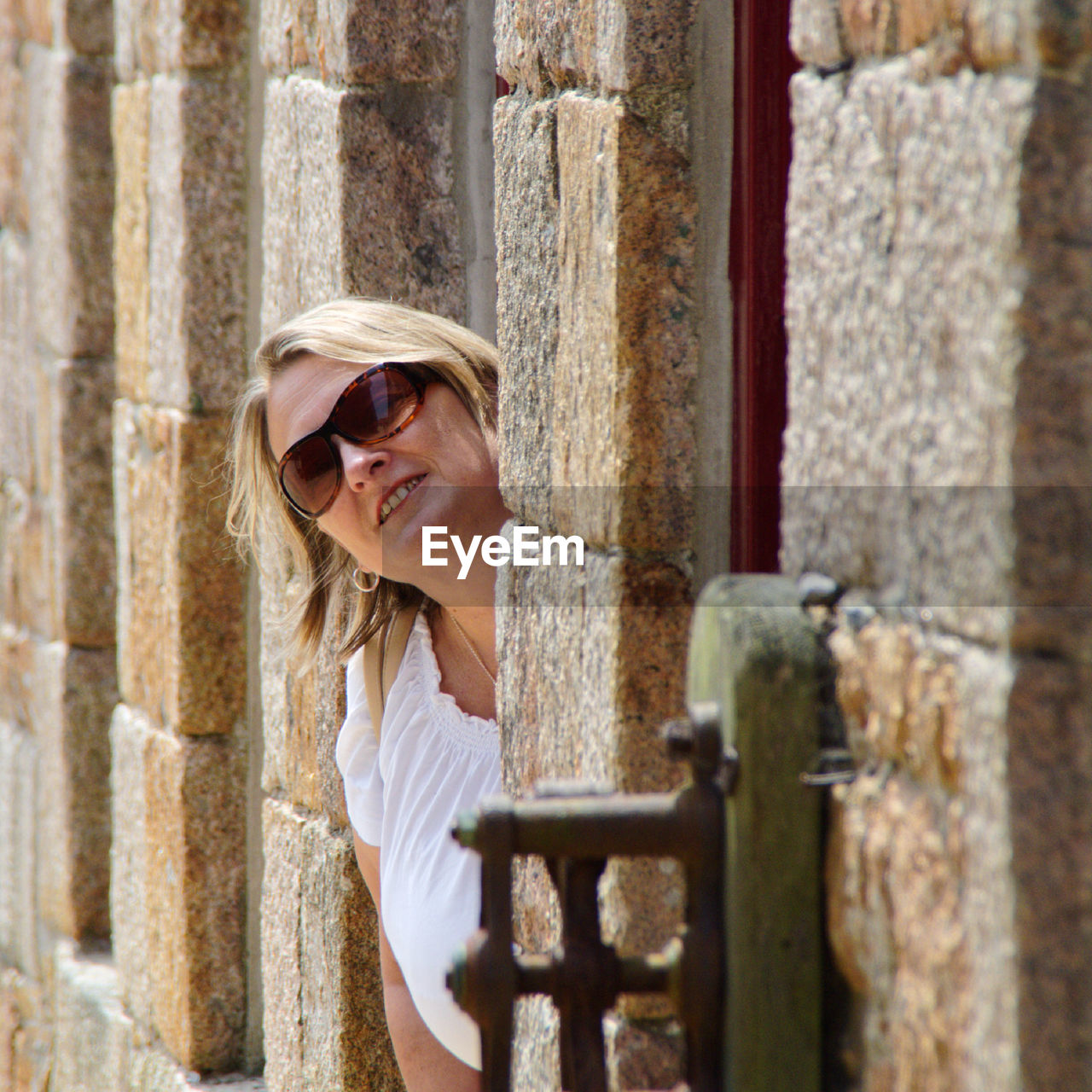 Portrait of woman wearing sunglasses while peeking through window on stone wall