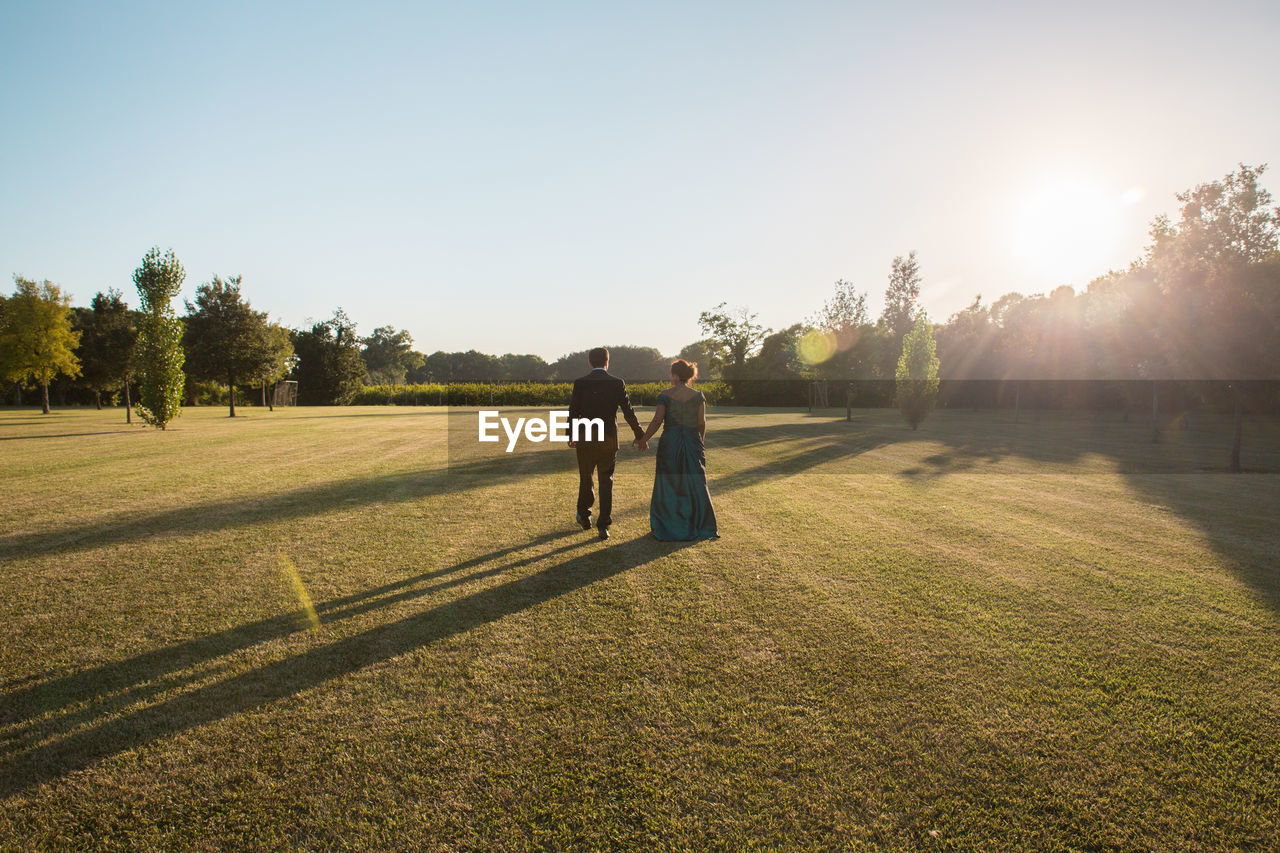 Rear view of couple holding hands while walking on grassy field against clear sky during sunny day
