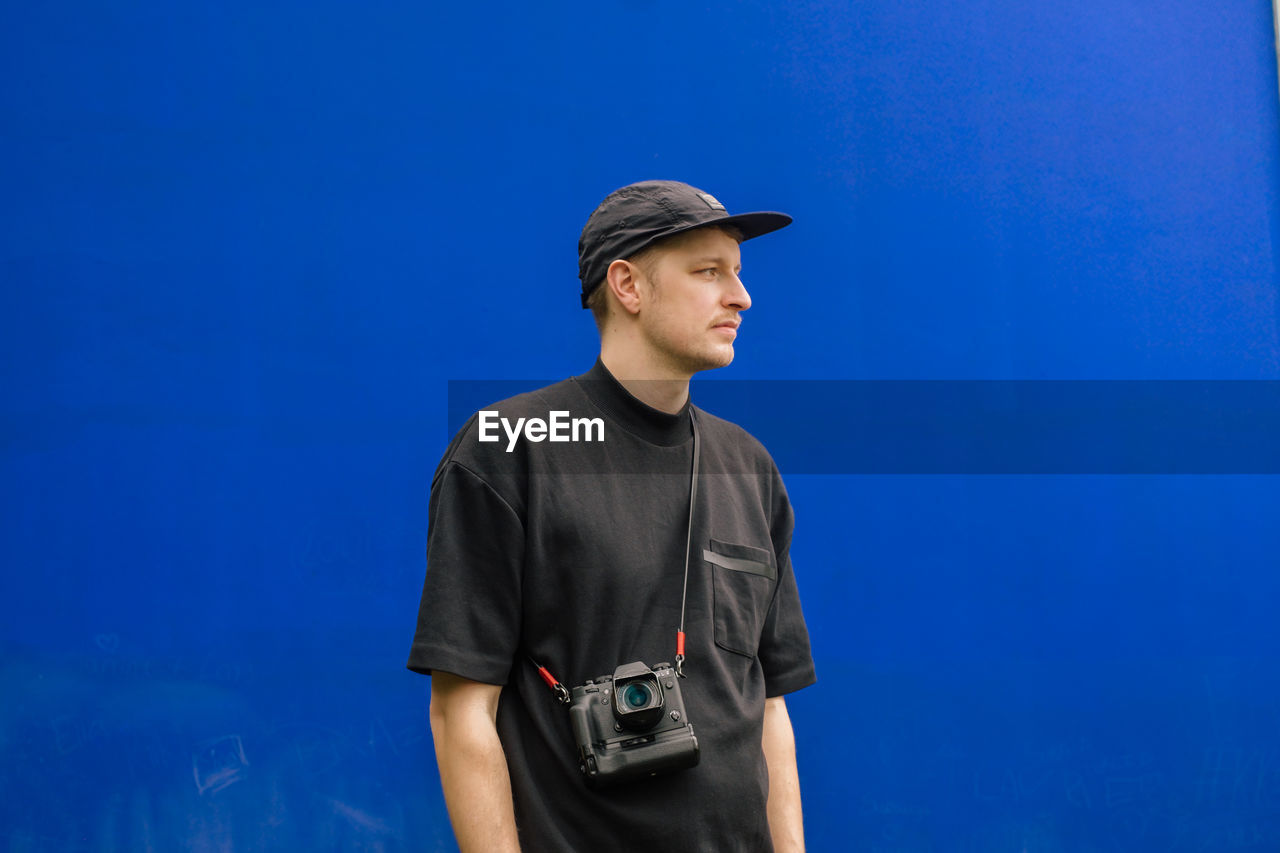 Low angle view of young man wearing cap looking away while standing against blue wall