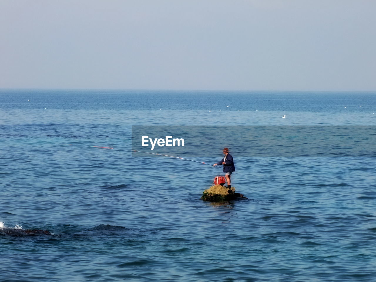 Side view of man fishing in sea while standing on road against clear sky