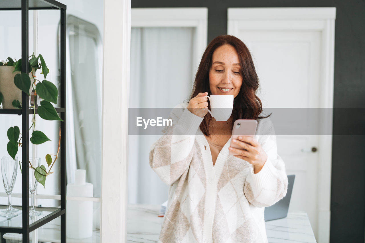 Smiling brunette woman with long hair in white cardigan using mobile phone in office