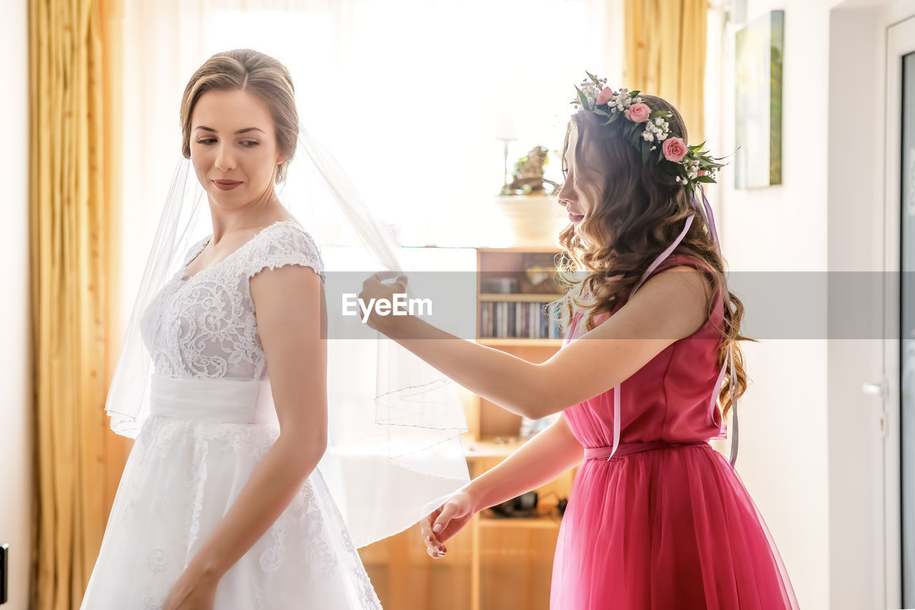 Side view of woman assisting bride getting dressed at home