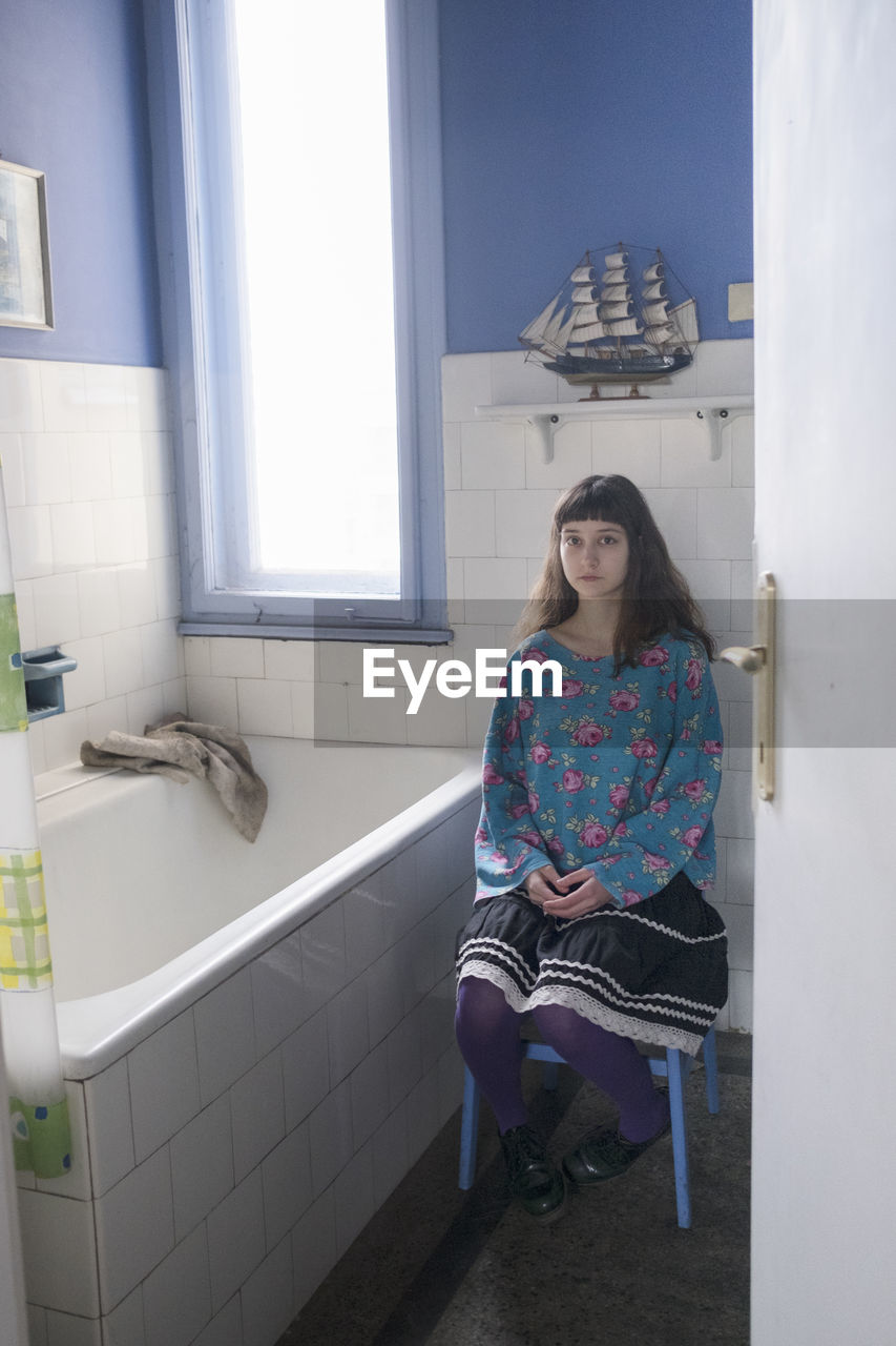 Portrait of young woman sitting in bathroom at home seen through doorway