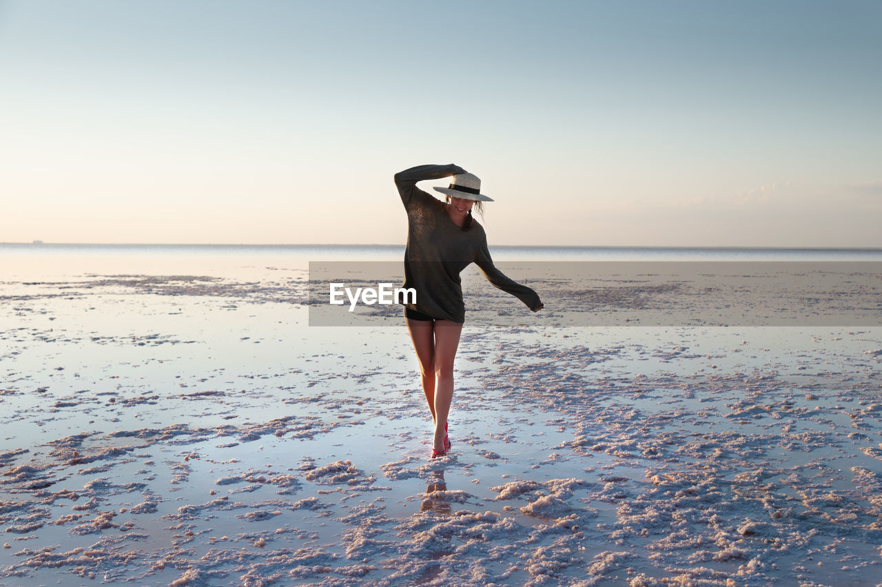 Attractive young caucasian woman in a straw hat and long jacket walks balancing on a shallow salt 