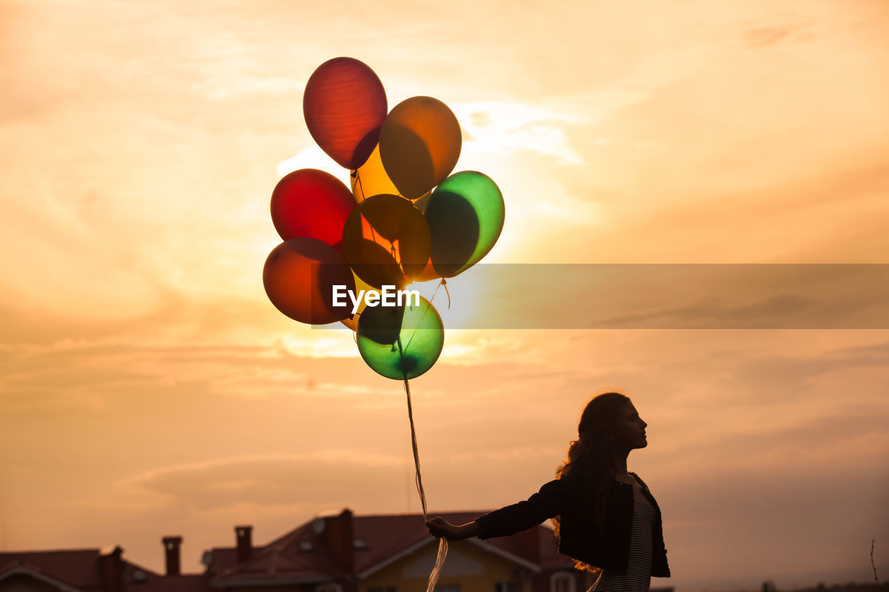 Silhouette of person holding balloons against sky during sunset