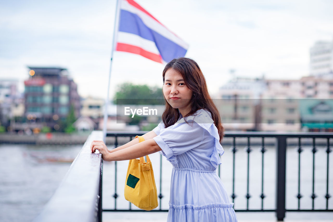 Portrait of woman standing by railing against flag and buildings in city