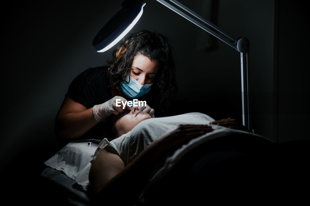 Professional cosmetician in medical mask cleaning face of female client with wet napkin while preparing for procedure of eyelash lifting in salon