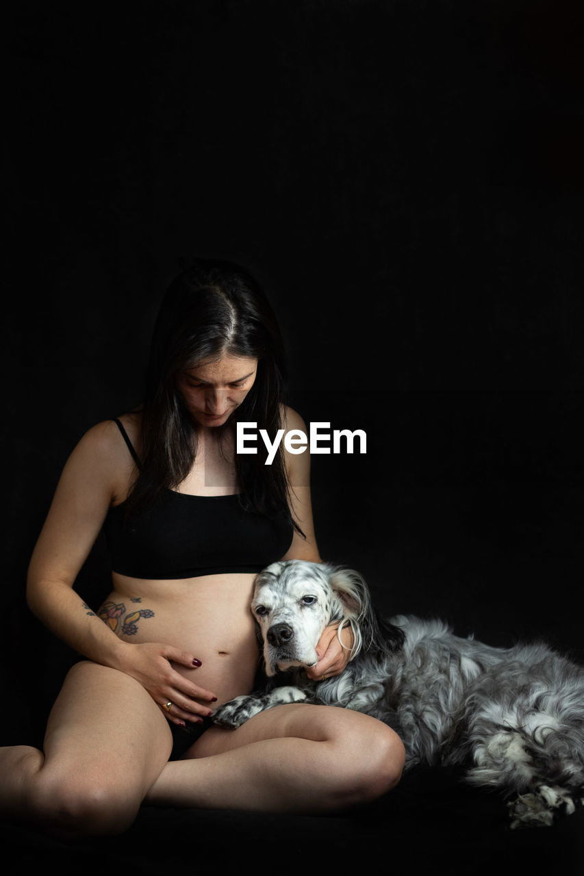 Portrait of pregnant woman with dog sitting against black background