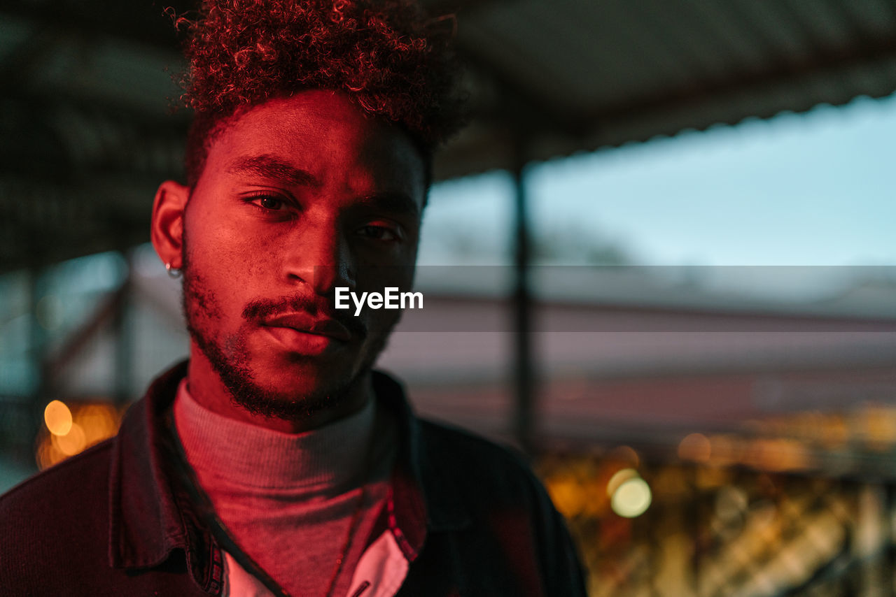 Young african american hipster male with curly hair and beard wearing warm jacket looking at camera while standing against shabby wall