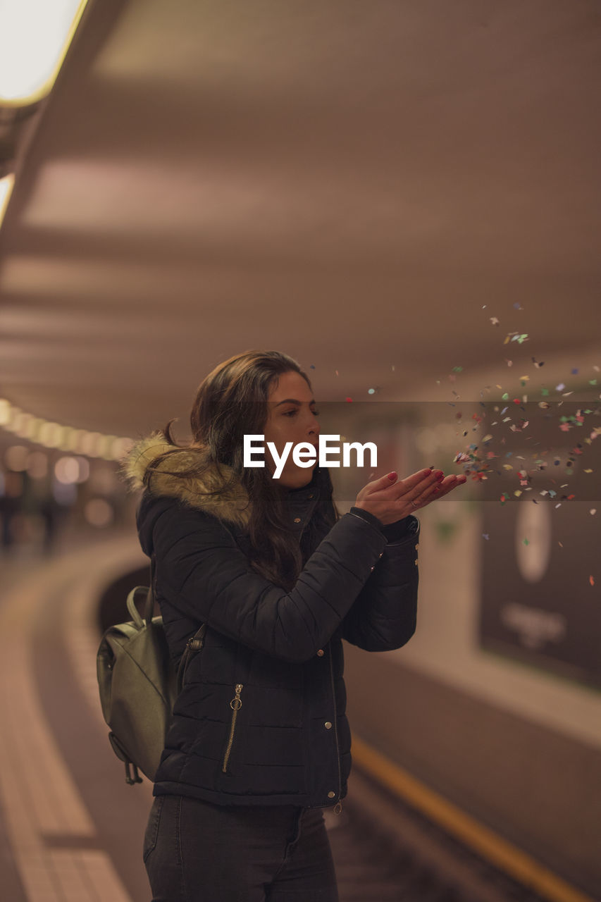 Young woman blowing confetti while standing against train at subway station