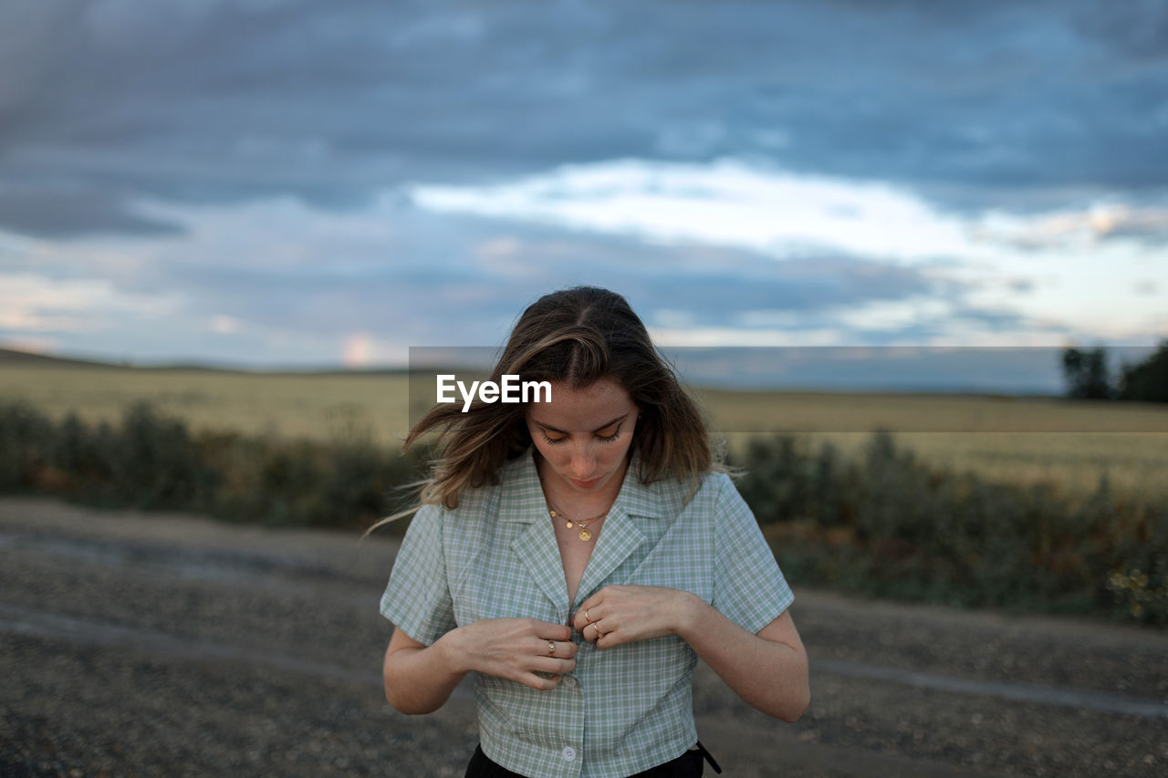 Trendy young female buttoning up shirt on roadway against field under cloudy sky in twilight