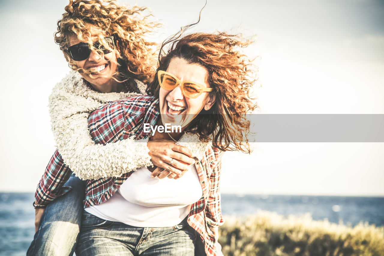 Smiling woman piggybacking friend against sky