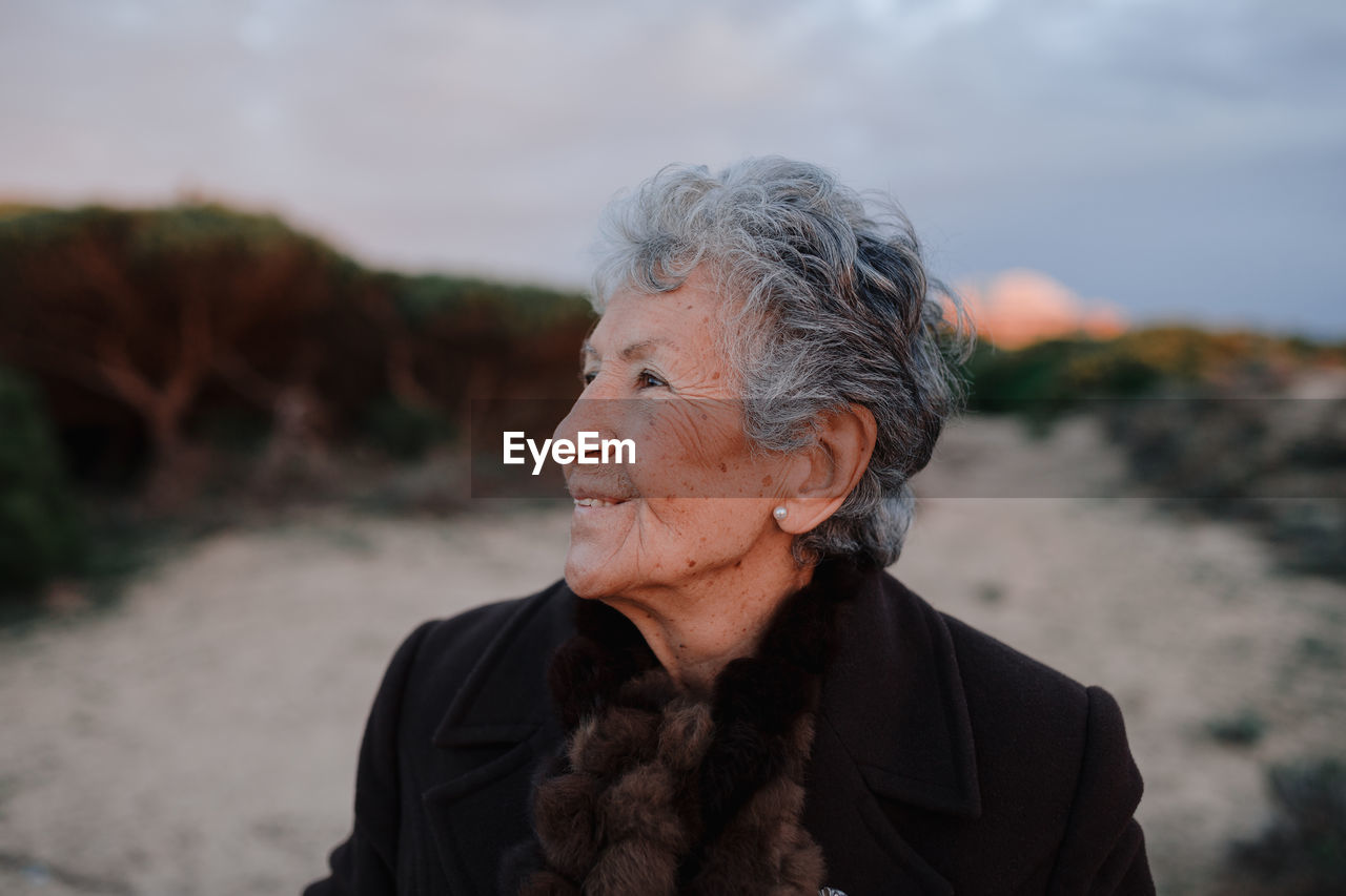 Happy elderly female tourist with gray hair in warm casual outfit smiling and looking away while relaxing on sandy beach against cloudy evening sky