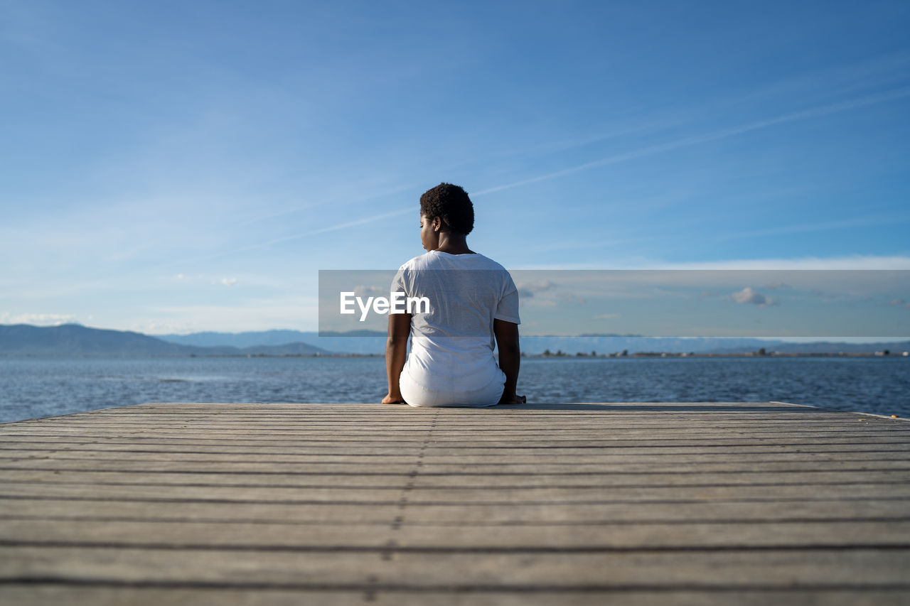 Back view anonymous african american female in white clothes sitting on wooden quay near vast rippling lake under clear blue sky in summer nature