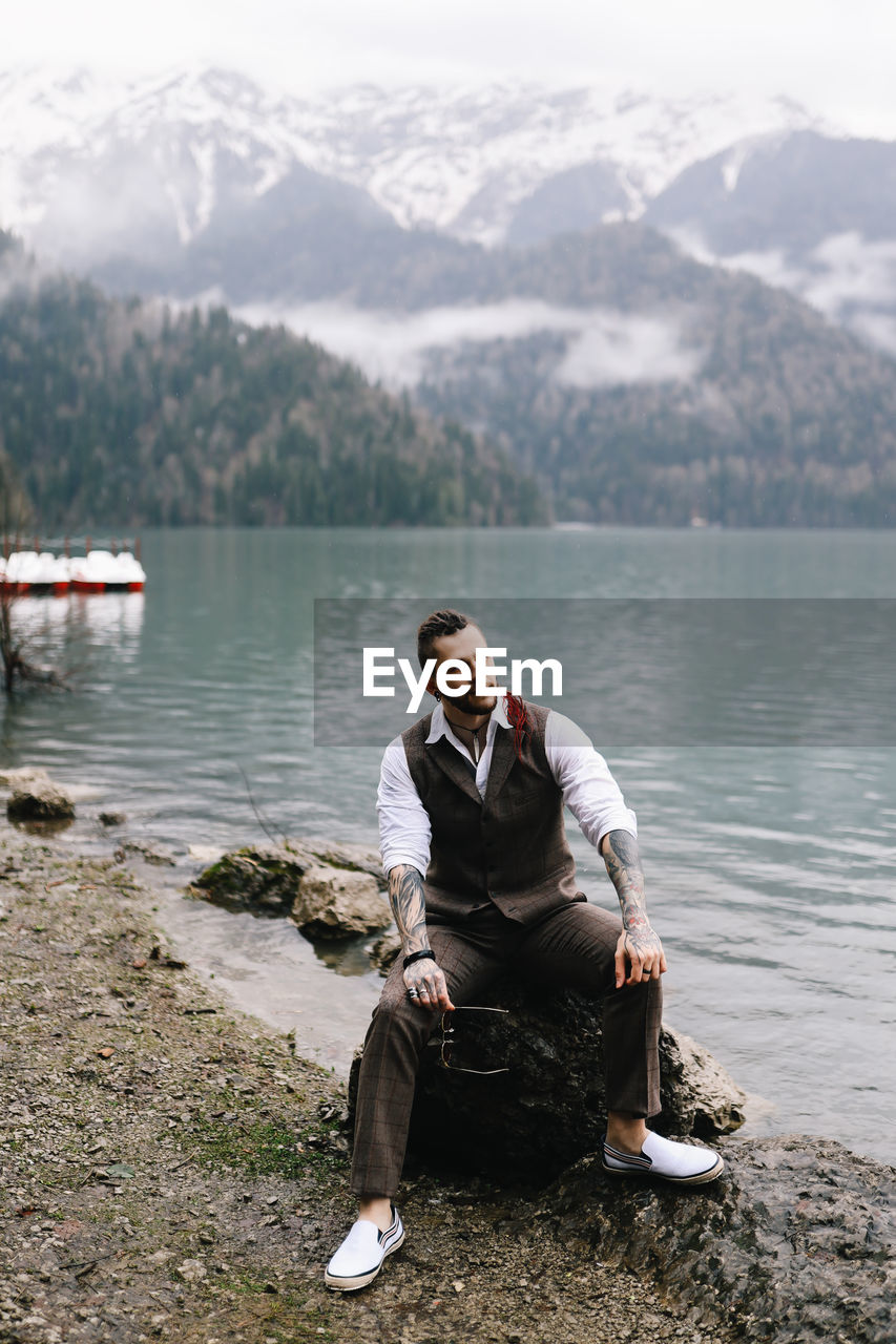 A tall brutal man hipster groom in a wedding suit stands by the misty mountains and lake in nature