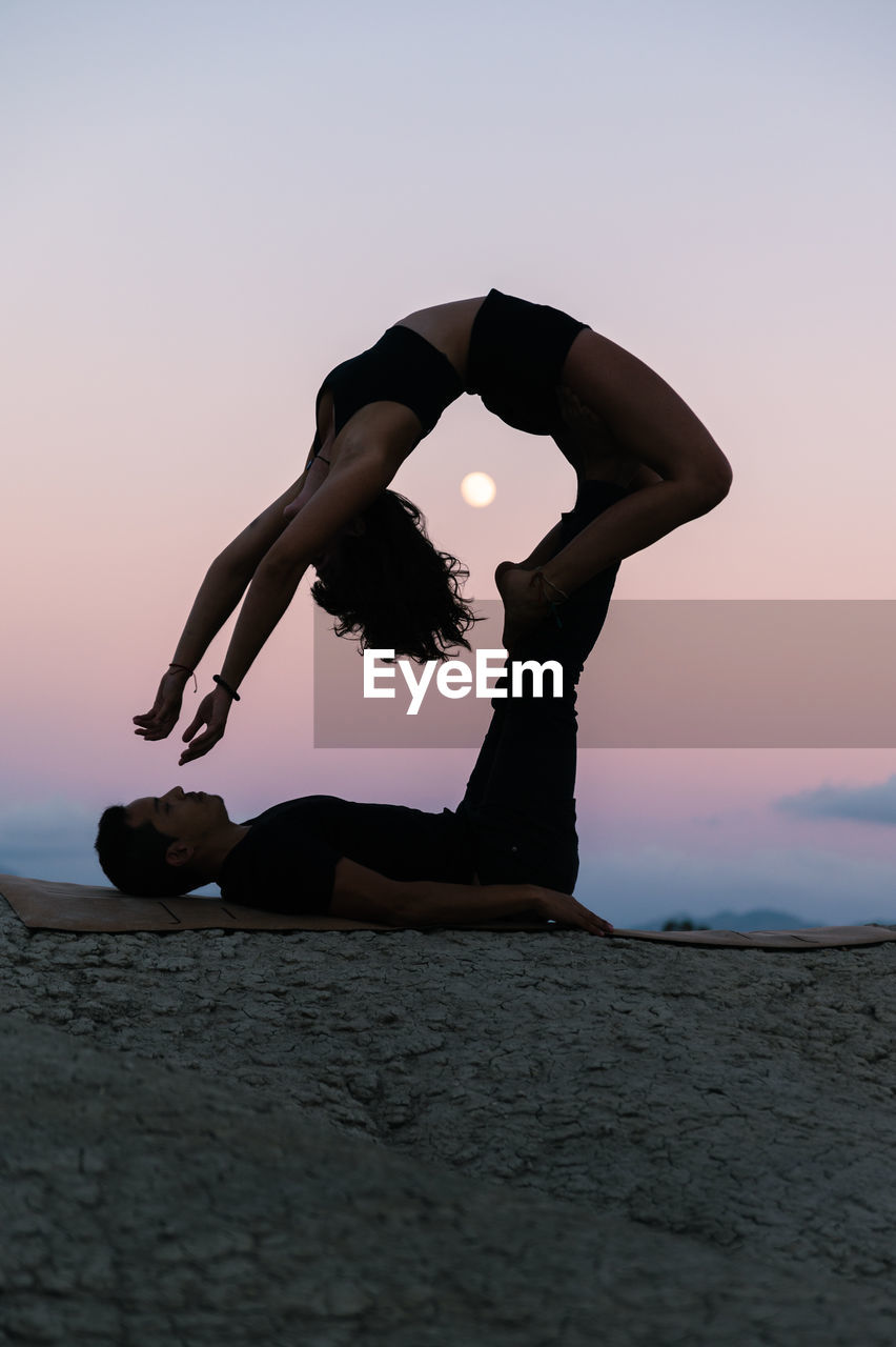 Side view of silhouette of flexible woman doing backbend and balancing on legs of man during acroyoga session against sunset sky with moon