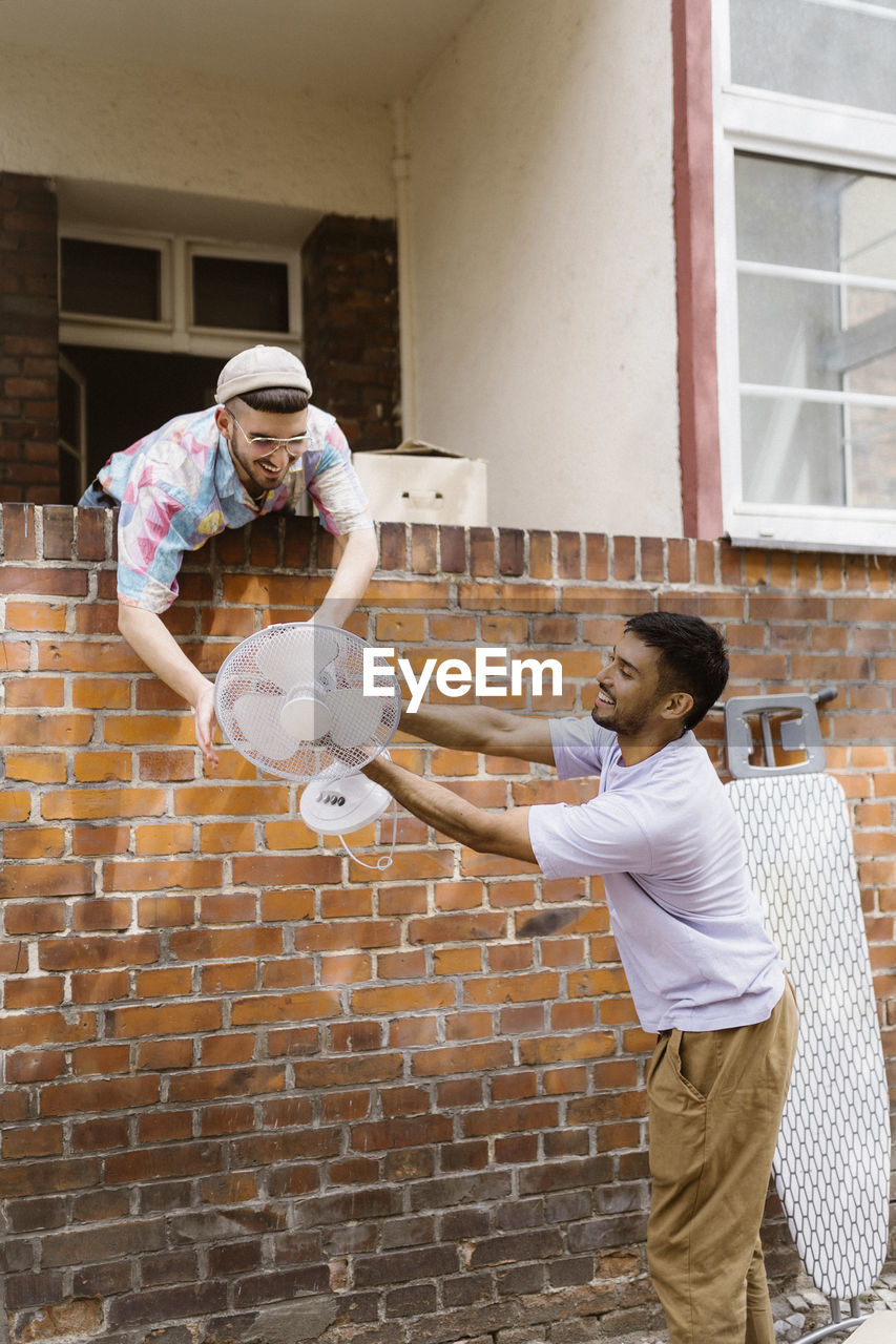 Smiling gay couple helping each other while holding electric fan over brick wall