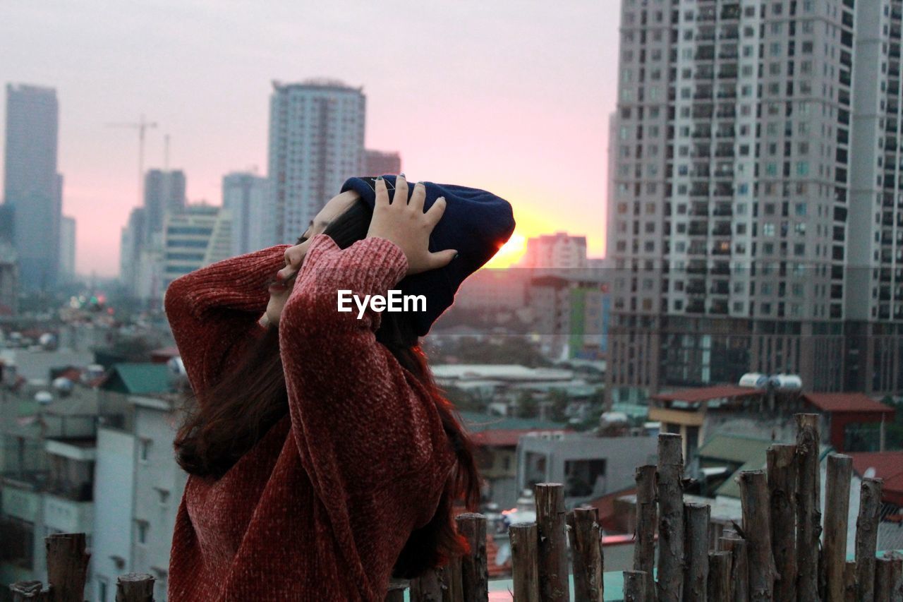 Side view of teenage girl with head in hands standing against buildings during sunset