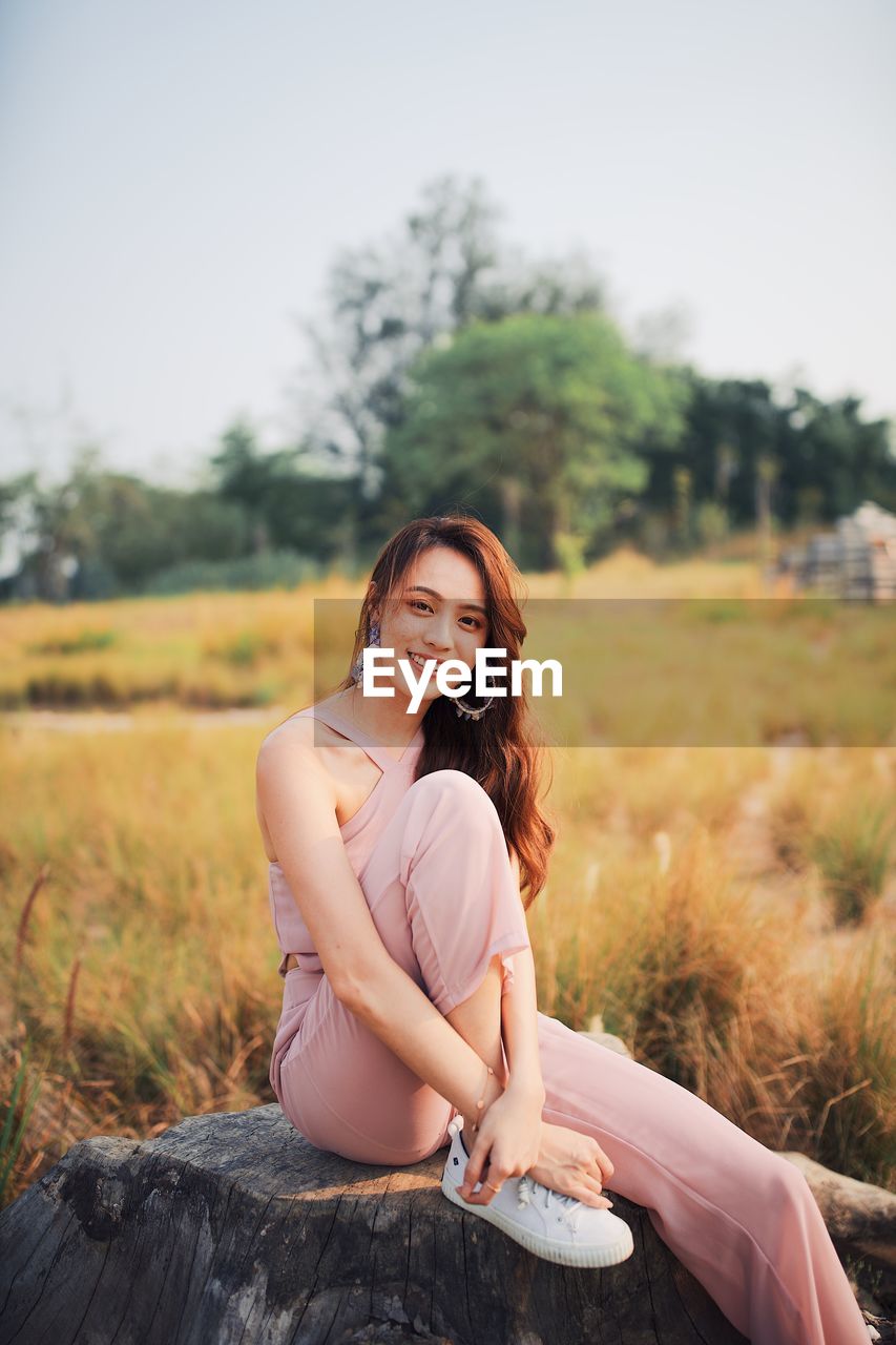 Portrait of young woman sitting on tree stump against clear sky