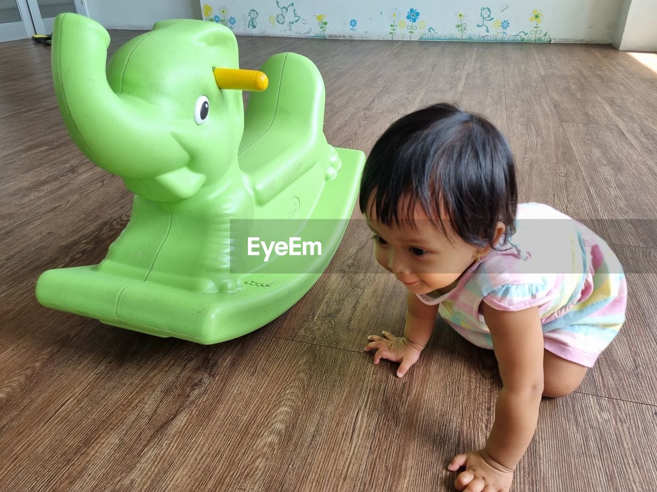 childhood, child, toy, flooring, one person, indoors, green, toddler, innocence, full length, hardwood floor, animal representation, person, baby, baby toys, cute, fun, high angle view, female, black hair, representation, home interior, lifestyles, wood, human head, happiness, domestic life, casual clothing, looking