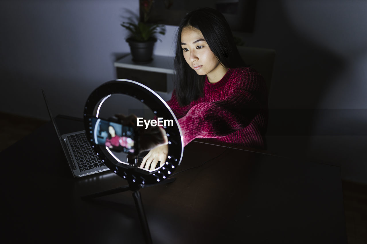 Young chinese female vlogger sitting with laptop and illuminated ring light