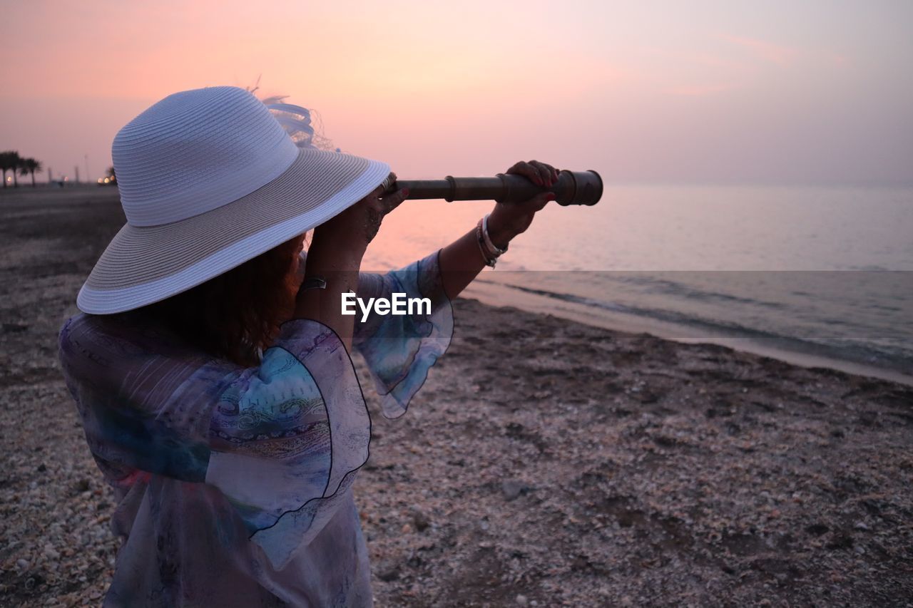 Woman looking through hand-held telescope at beach against sky during sunset