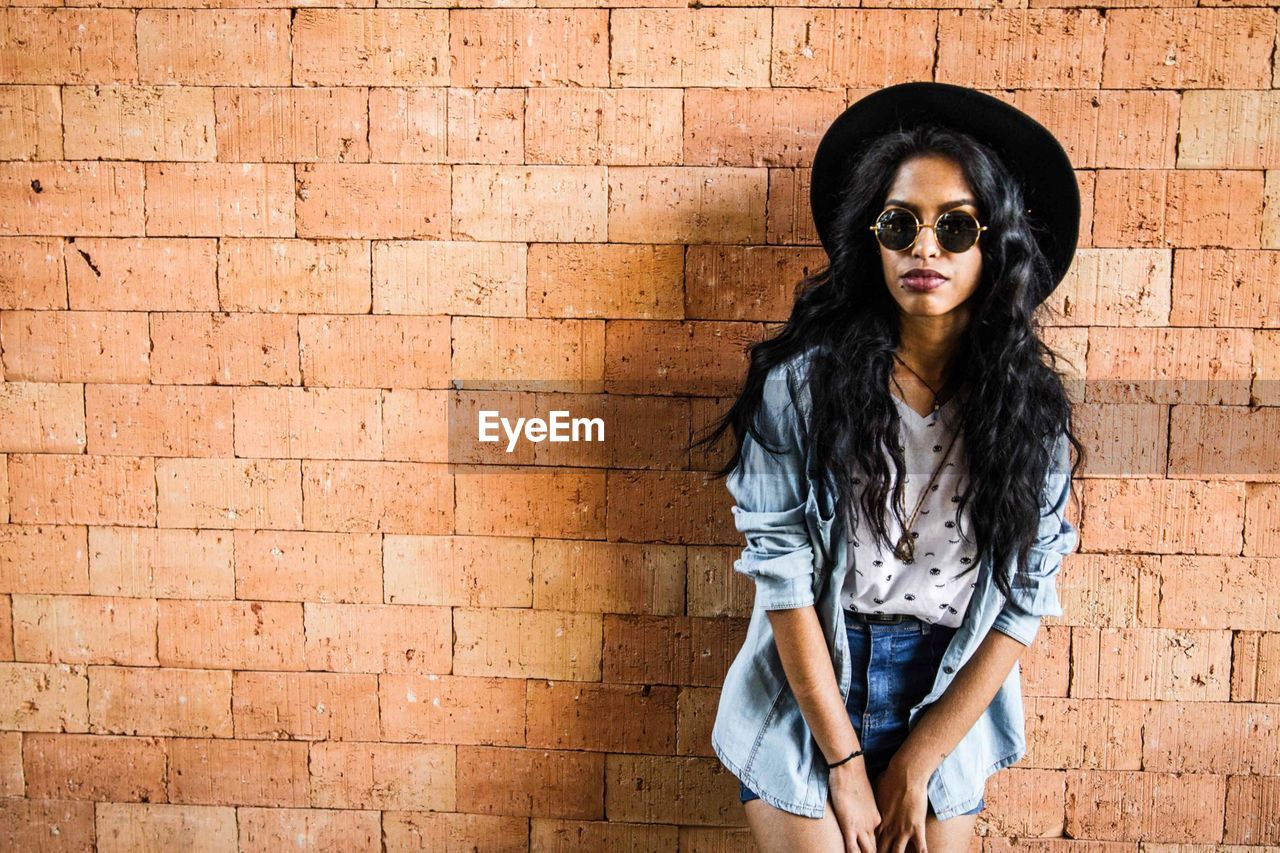 Portrait of young woman wearing sunglasses standing against brick wall