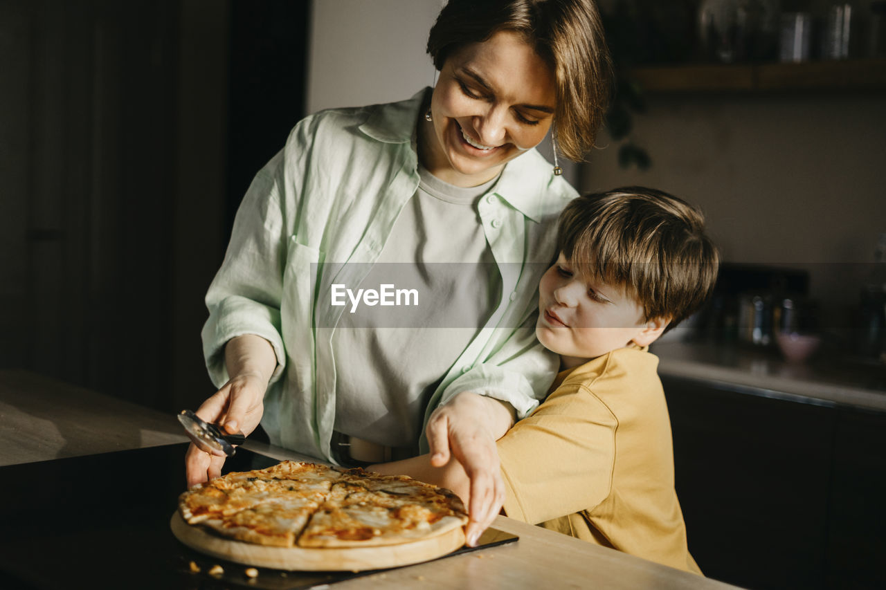 Son embracing mother holding pizza and cutter at home