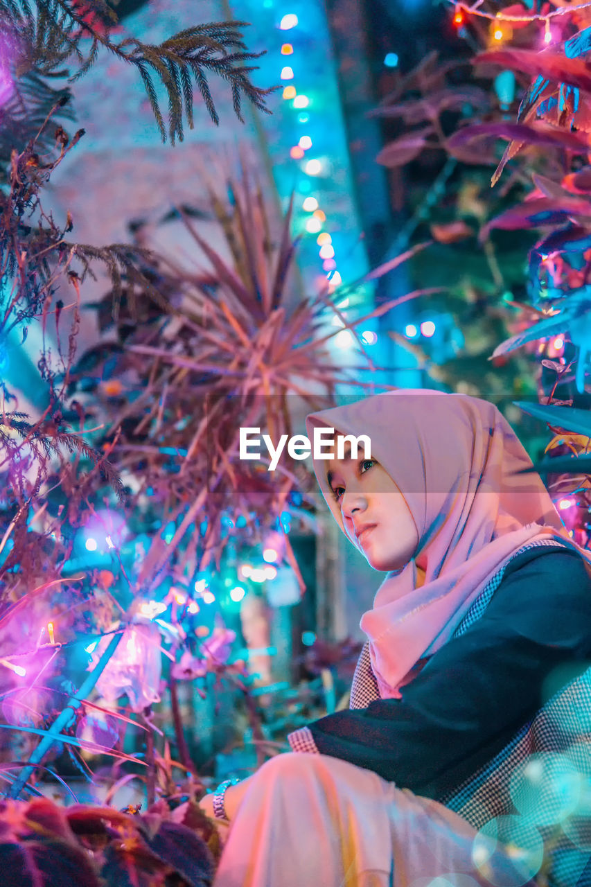 Woman wearing hijab while sitting amidst illuminated plants