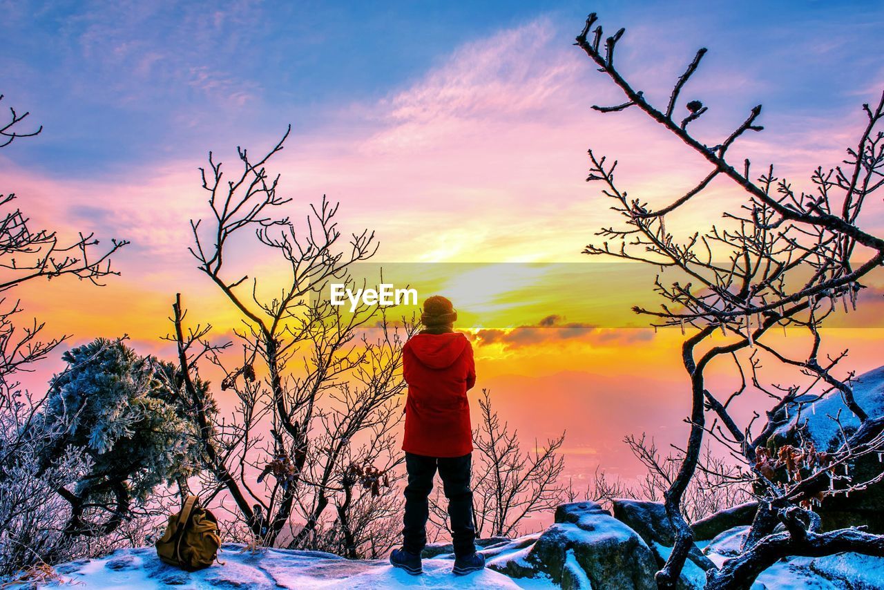 Rear view of mid adult man standing on snow covered mountain against sky during sunset