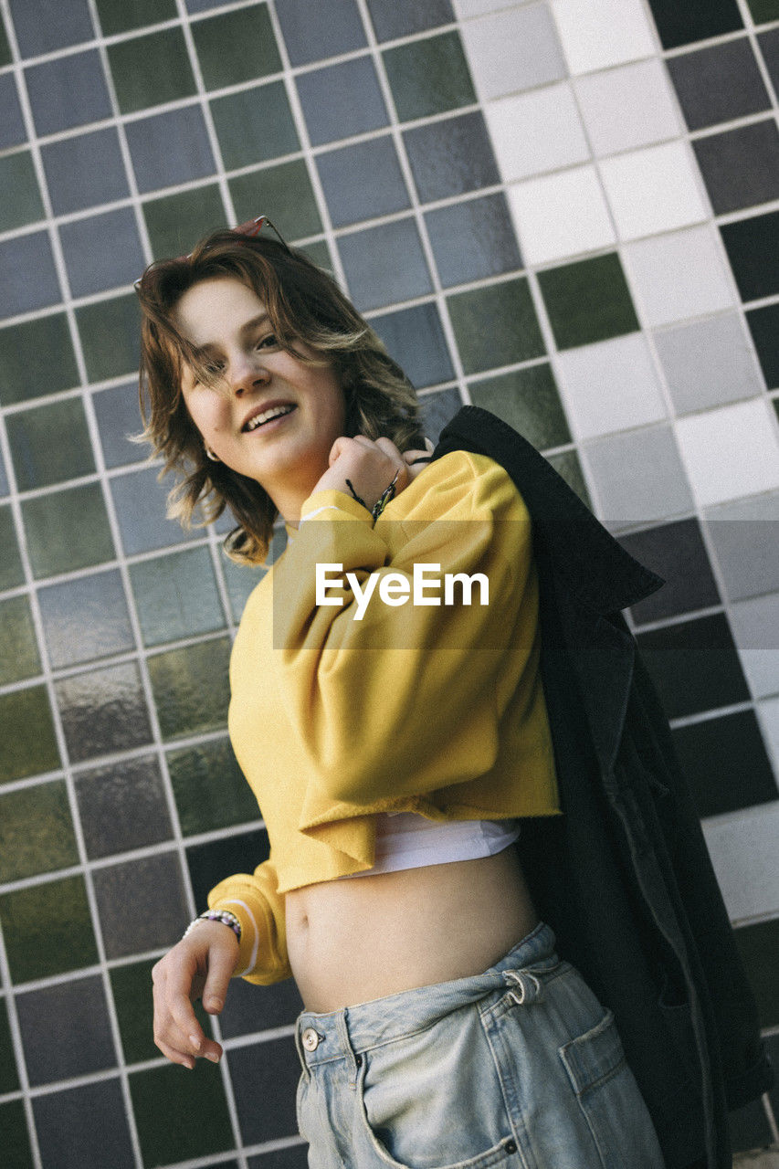 Portrait of smiling young woman holding jacket while standing against tiled wall