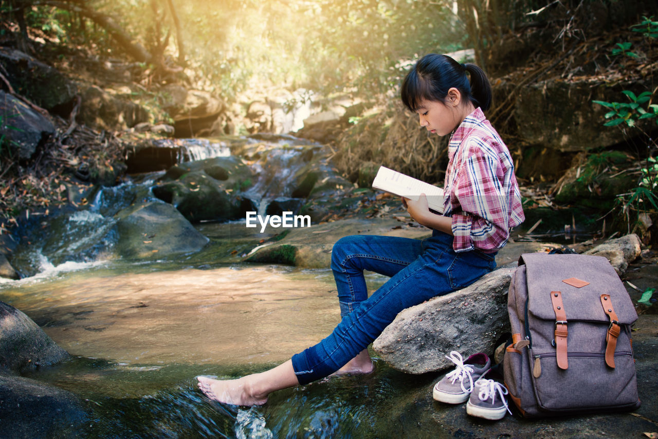 Girl reading book while sitting on rock at river