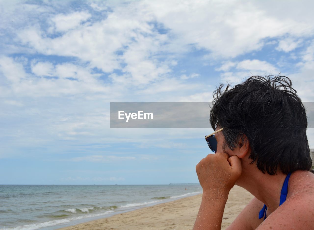 Side view of woman sitting on beach against cloudy sky