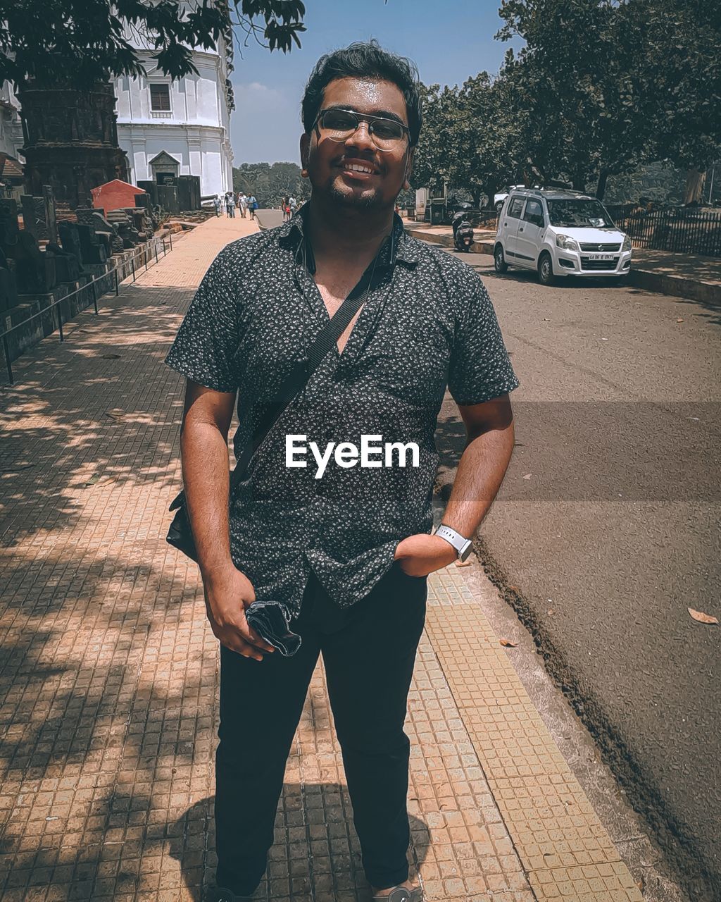 one person, adult, portrait, front view, looking at camera, men, glasses, standing, transportation, fashion, casual clothing, car, sunglasses, city, lifestyles, young adult, smiling, vehicle, motor vehicle, architecture, day, facial hair, beard, black, street, nature, mode of transportation, happiness, sunlight, outdoors, clothing, road, emotion, person