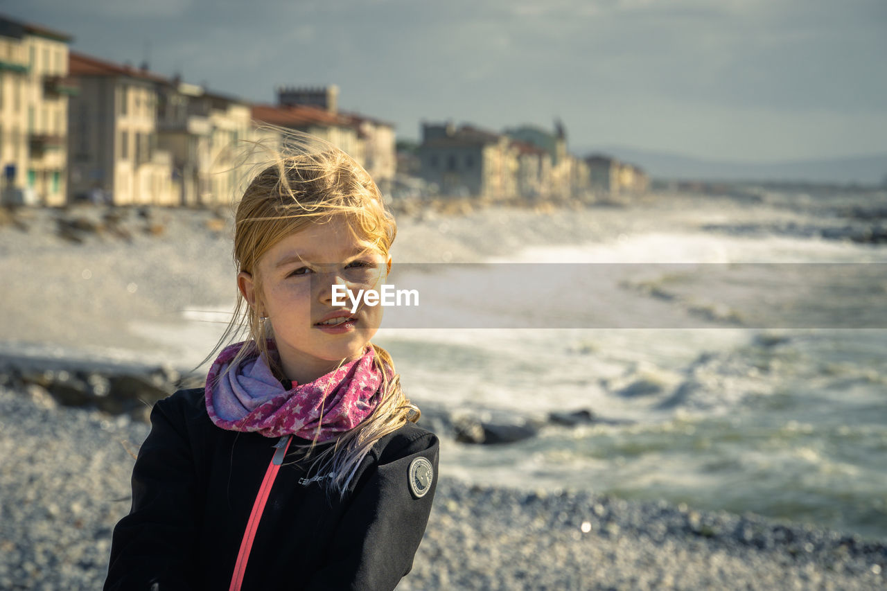 Girl posing in front waterfront against sky, breaking waves on rocky beach, marina de pisa