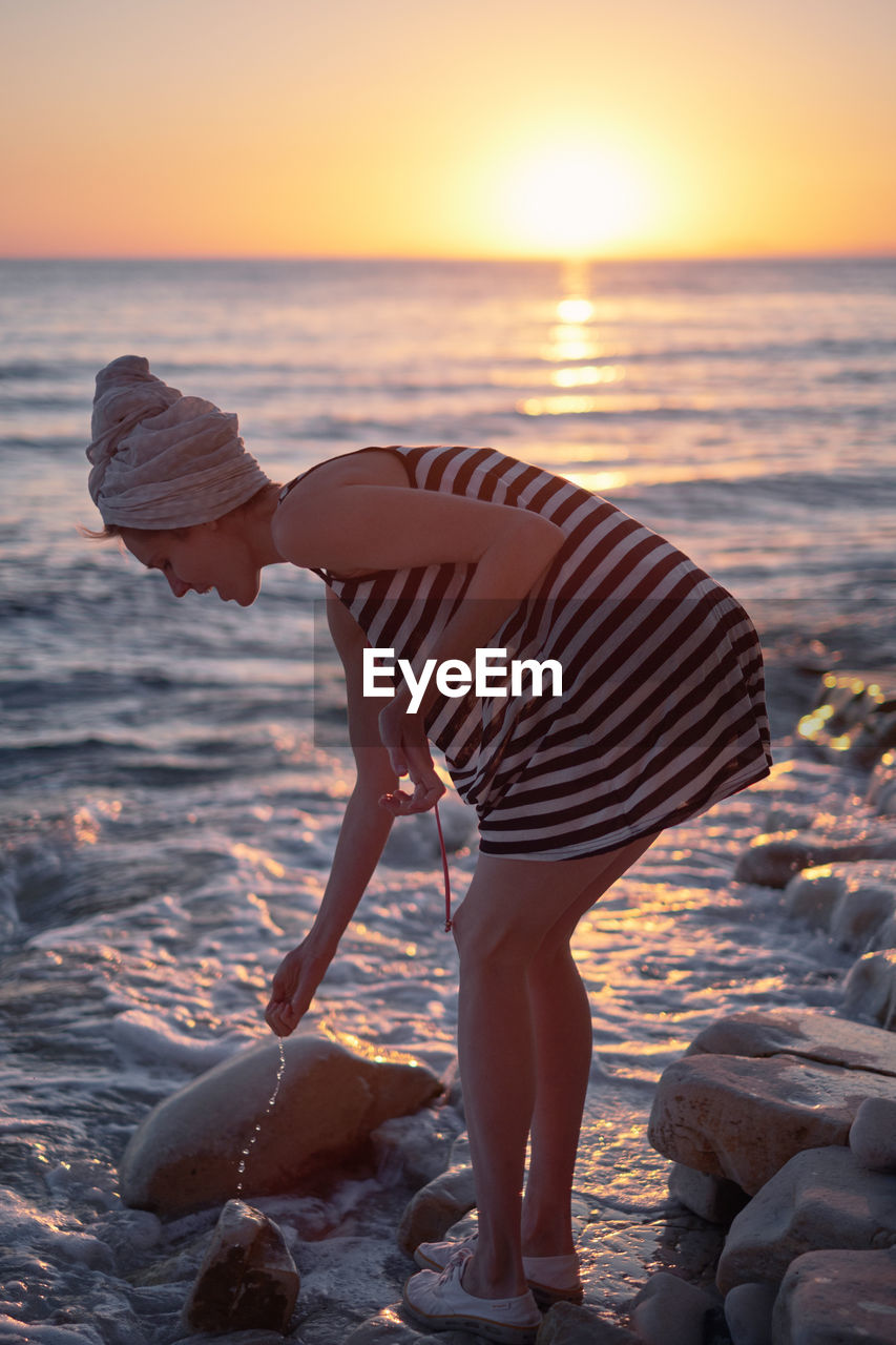 Full length of woman on rock at beach against sky during sunset