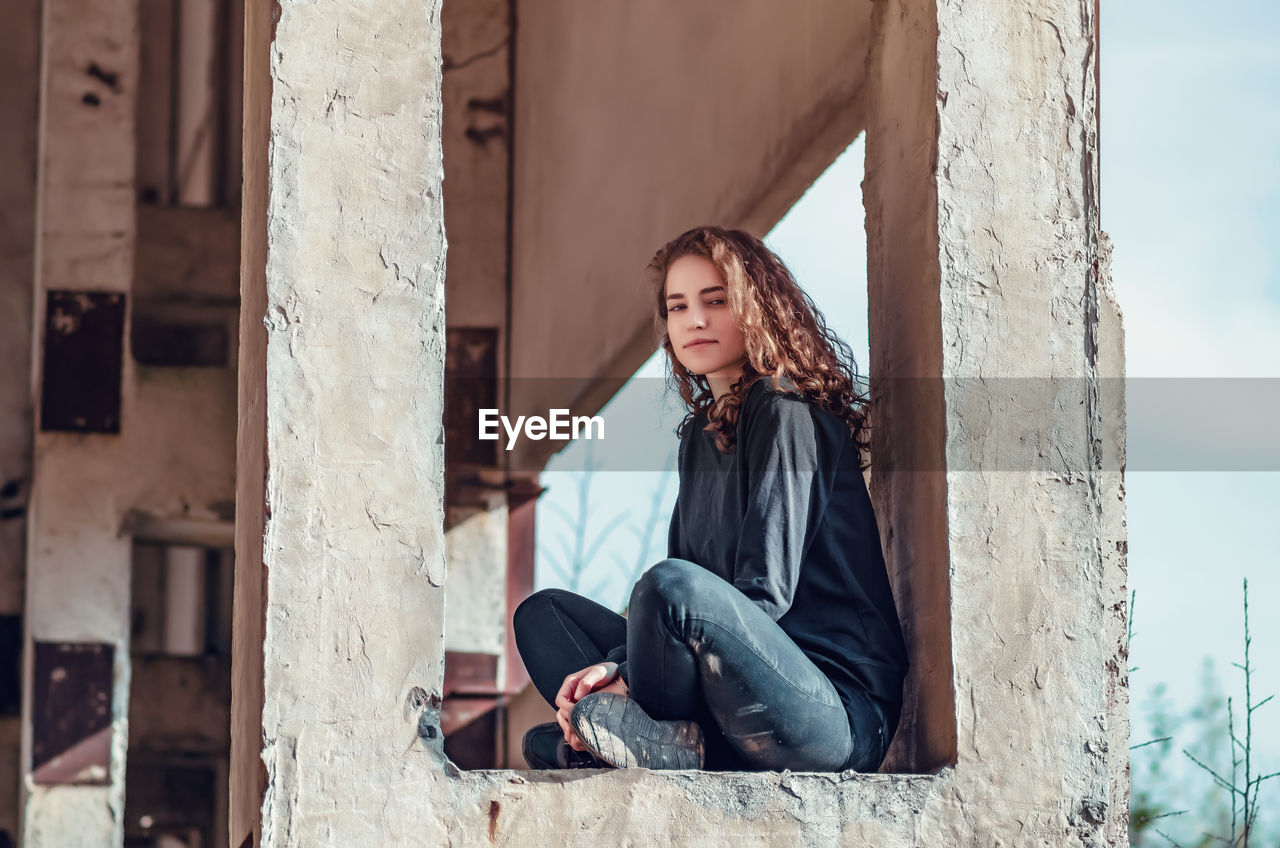 Curly cute young woman sitting in window of unfinished house. architecture, construction.