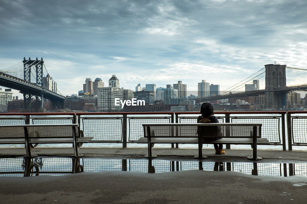 Rear view of women sitting on bench against brooklyn bridge over east river