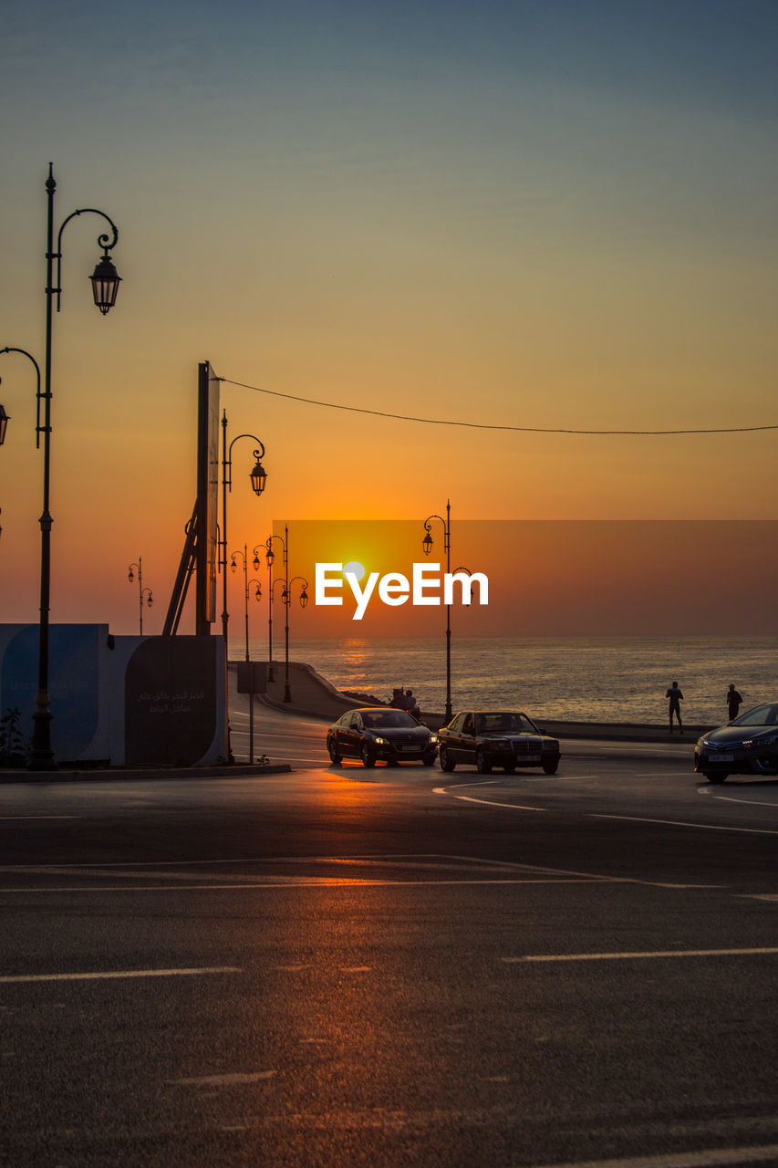 Cars on street by sea against sky during sunset