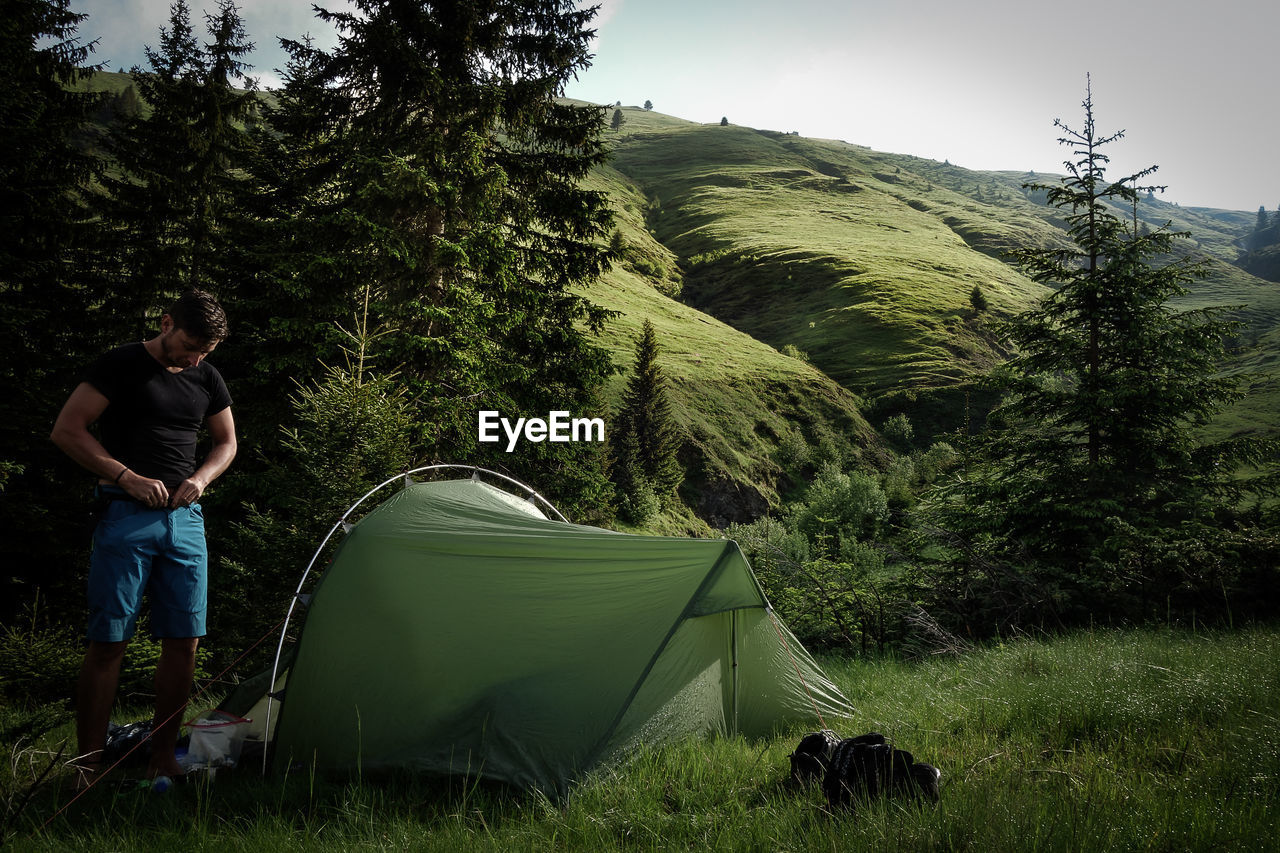 Man fastening shorts while standing by tent on field against sky