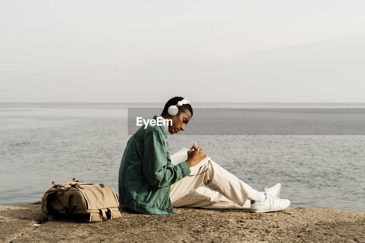 Young man with headphones writing in diary by sea