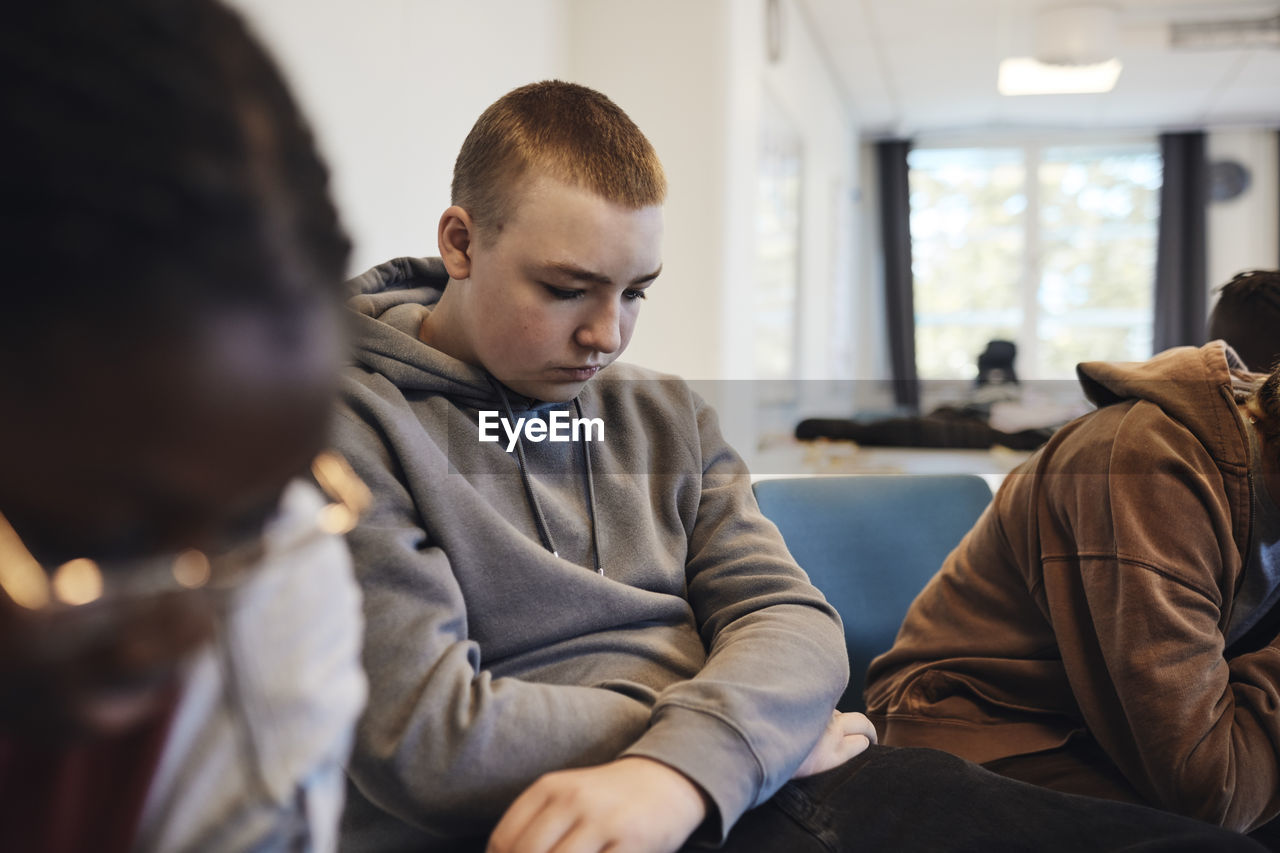 Depressed teenage boy with short hair sitting amidst female friends in group therapy at school