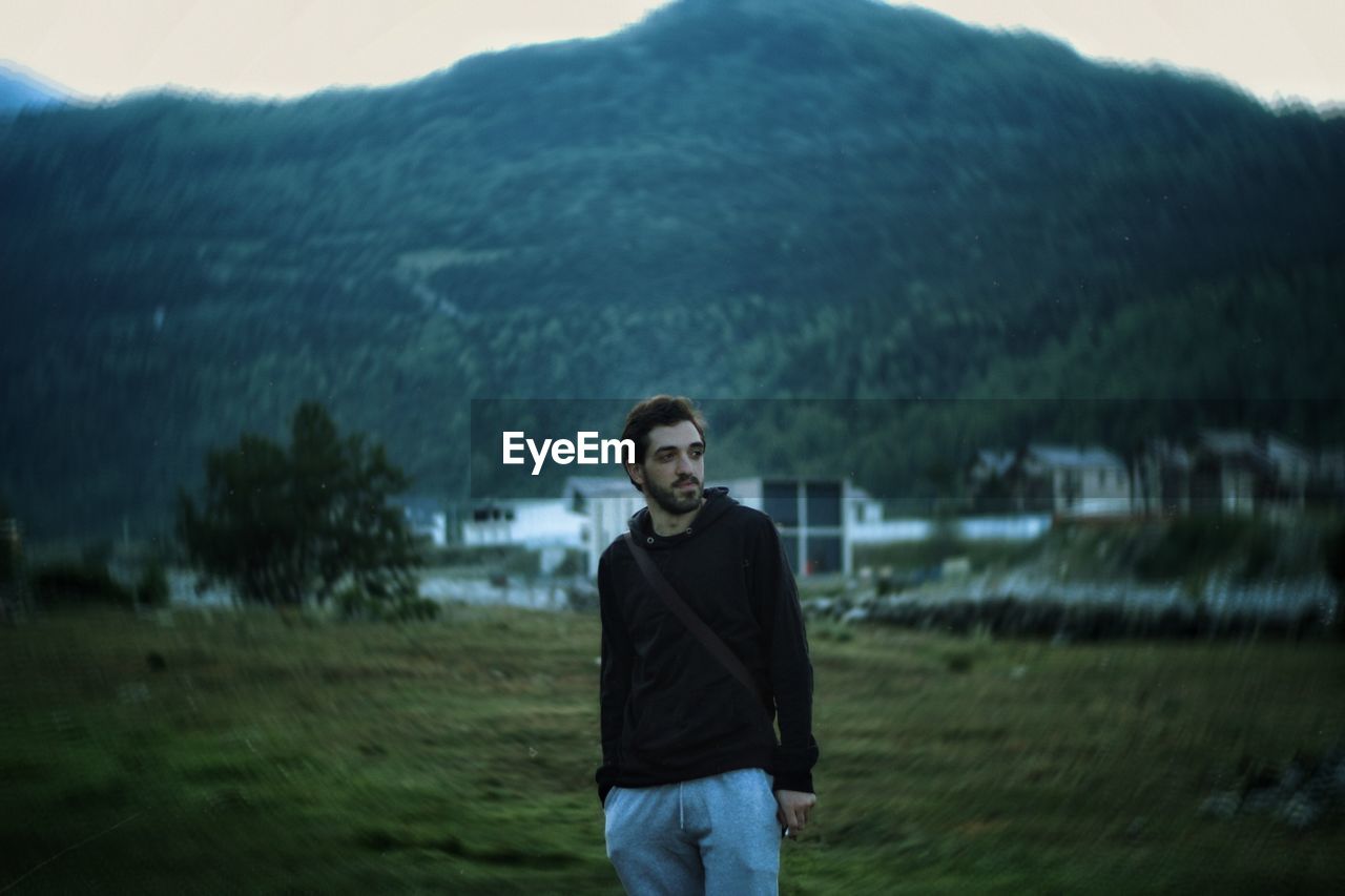 PORTRAIT OF YOUNG MAN STANDING ON FIELD AGAINST MOUNTAINS