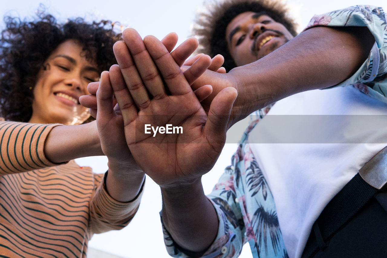 From below of positive young african american male and female putting hands together and smiling while having fun on sunny day